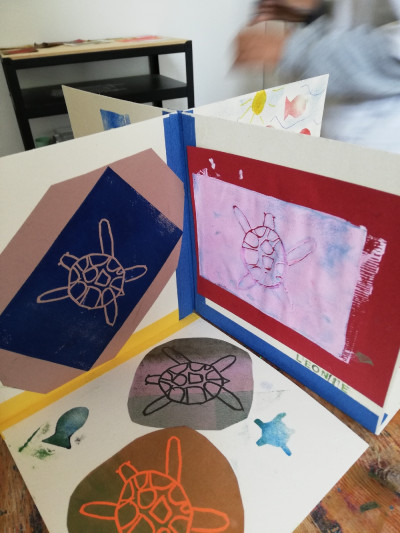 Ein Buch mit bunten Drucken von Schildkröten ist zu sehen, es ist aufegklappt auf einem Tisch und sieht von oben aus wie ein Kreuz mit verschiedenen Seiten.