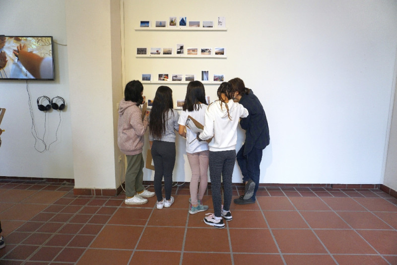 Vier Schüler:innen stehen vor einem Regal mit Fotografien, eine weibliche Person erzählt etwas dazu.