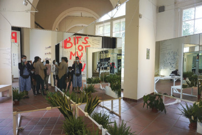 Zentral im Bild ist ein großer Spiegel in der man eine Gruppe von Kindern und Erwachsenen sieht. Vor dem Spiegel stehen Pflanzen auf dem Boden.