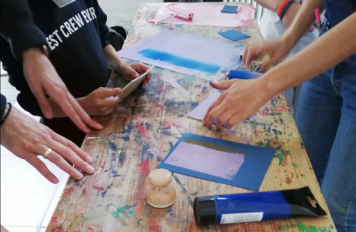 Sechs Hände auf einer Werkbank, sie halten Druckvorlagen und färben sie blau und rosa ein.