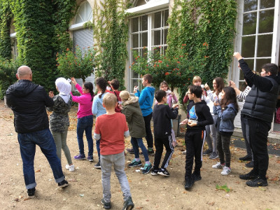 Ein Gruppe Schüler stehen vor einem Gebäude mit Efeubewuchs und schauen dabei alle etwas in der hochgehobenen Wand an.