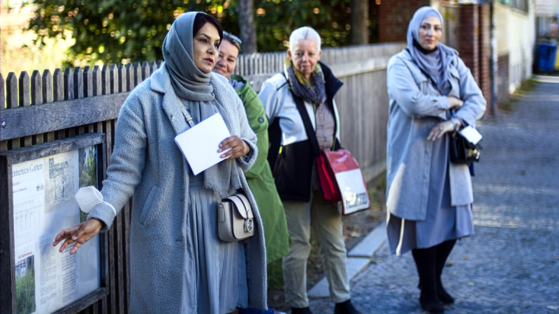 Eine Frau steht vor einem Holzzaun und zeigt auf ein Schild, drei Frauen stehen recht neben ihr im Hintergrund