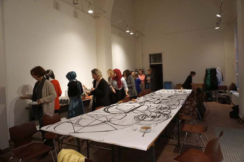 Ein langer Tisch ist mit Papier bedeckt, darauf sind große schwarze Linien von Tuschezeichnungnen zu sehen, im Hintergrund eine Reihe Frauen, die sich Essen von einem Tisch nimmt.