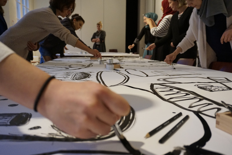 Im Vordergrund ist eine Hand, die mit einem Pinsel ein große schwarze Tuschezeichnung auf weißes Papier malt, im Hintergrund sind weitere Person, die das Gleiche tun.