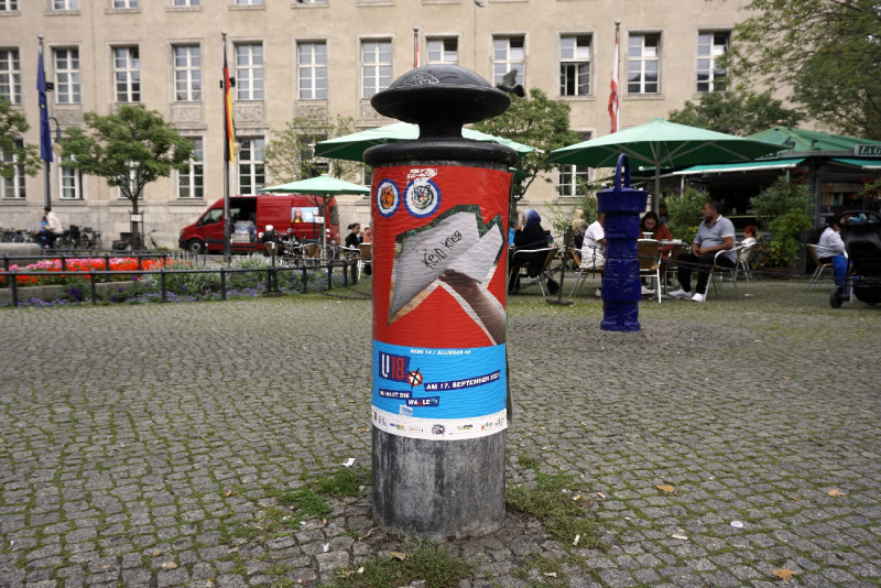 Buntes Plakat auf Hydrantem, Platz mit Cafe ist im Hintergrund zu sehen