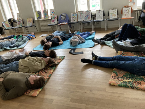 Schüler*innen liegen mit Augen zu auf dem Boden auf bunten Decken, um sie herum stehen Stühle mit gezeichneten bunten Bildern.