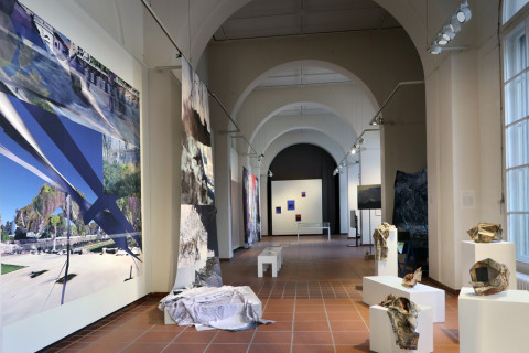 Blick in eine Ausstellung. Sie ist in einem langen Raum an dessen Seiten links und Rechts Bilder und Skulpturen zu sehen sind.