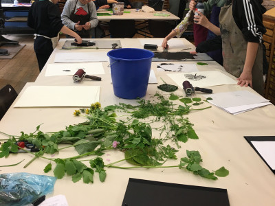 Auf Tischen liegen Pflanzenteile und weiße Papiere, Menschen stehen drum herum und färben Plexiglasplatten in schwarzer Farbe mit Linolrollern ein, die neben daneben liegen.