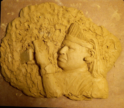 Aus einem Haufen Ton tritt das Relief eines Gesichtes hervor. Die Person hält ein Hnday vors Gesicht und fotografiert damit.