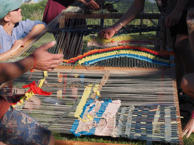 Ein großer Webstihl mit bunten Fäden bespannt, drum herum stehen Menschen und weben darauf