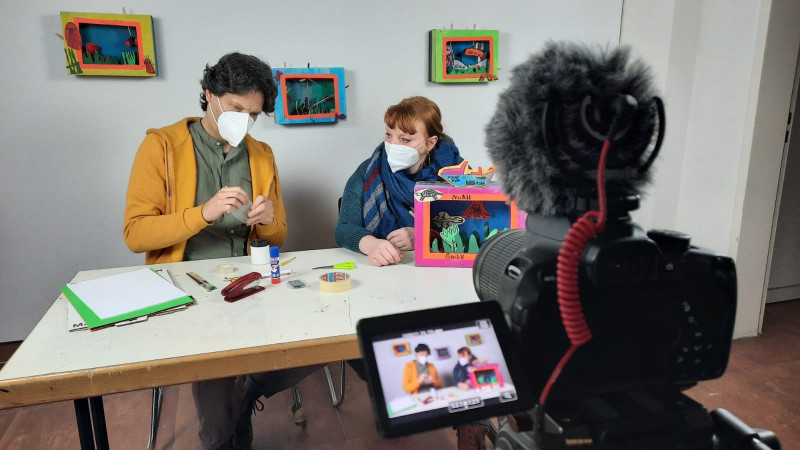 Ein Mann und eine Frau sitzen an einem weißen Tisch. Darauf liegen Bastelutensilien und ein bunte angemalte Pappkiste, in die ein kleine Landschaft eingebaut ist.