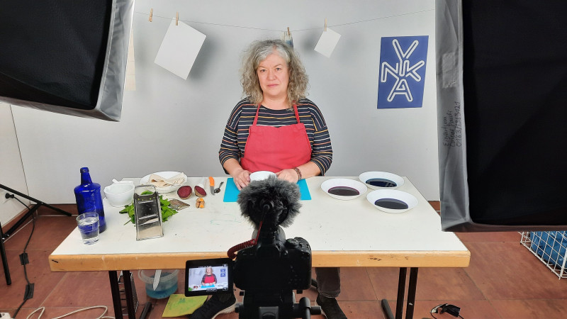 Eine Frau sitzt vor einem weißen Tisch. Darauf stehen drei Teller mit blauer Farbe recht und links eine blaue Glasflasche, ein Glas Wasser, Spinat und ein Hobel, rote Beete.