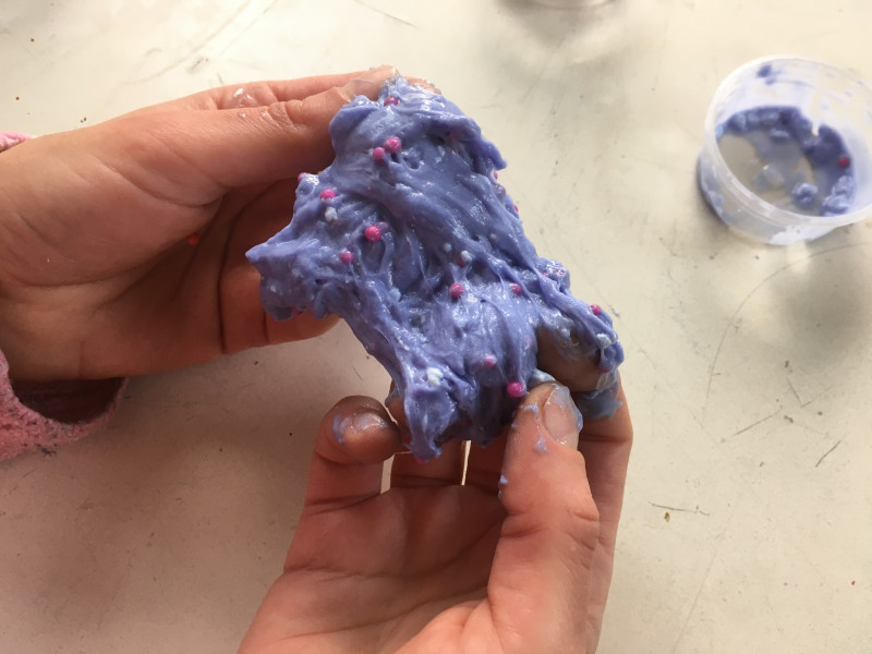 Zwei hände ziehen eine gummiartige, lila farbene Masse (sog. Slime) auseinander, bunte Kügelchen sind darin zu sehen.